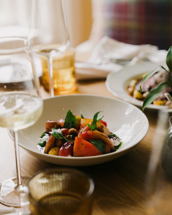 Fresh salad with tomatoes and herbs in a white bowl, surrounded by glasses and a blurred dining setting.
