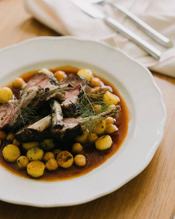 Rack of lamb on a white plate with potatoes, chickpeas, and sauce, garnished with herbs, on a wooden table.