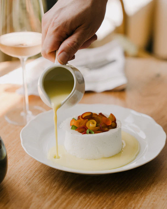Person pouring sauce over a dessert with fruit on a white plate.