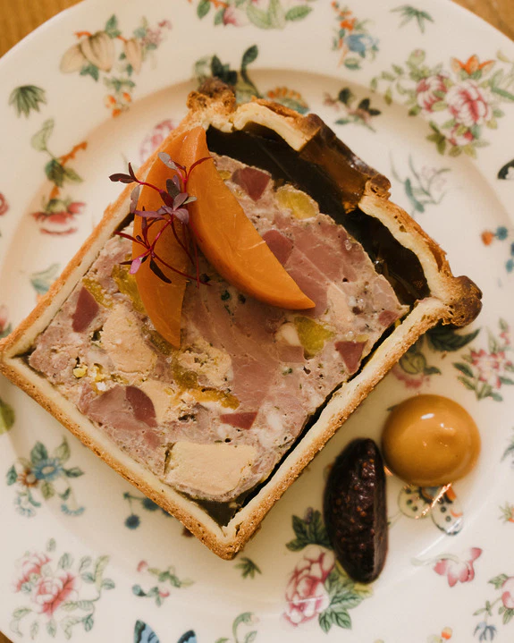 Slice of pâté en croûte with apricot garnish on a floral plate, served with accompanying sauces.