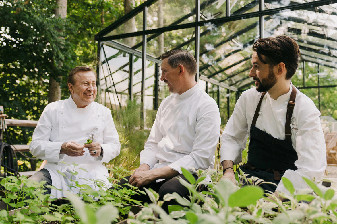 Three chefs in white coats sitting and chatting in a greenhouse, surrounded by greenery and plants.
