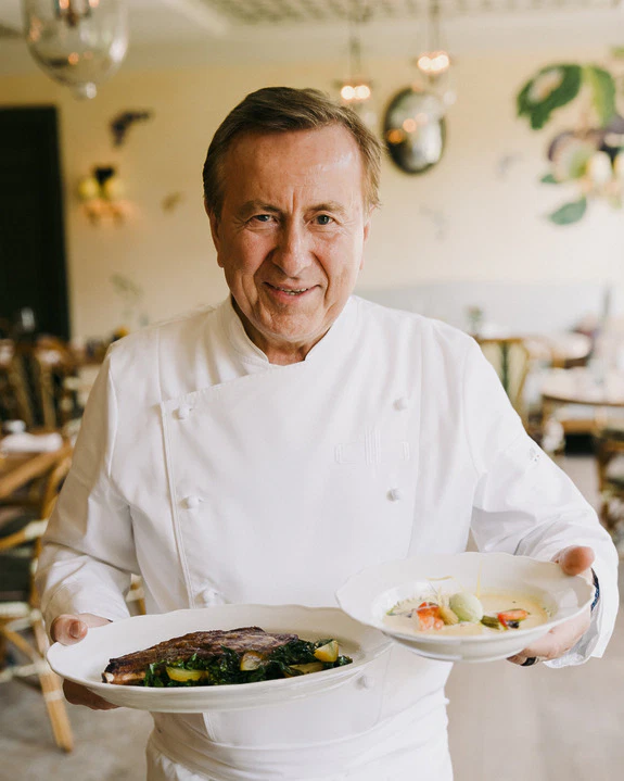 Chef in a white coat holding two plates of gourmet dishes inside a restaurant with elegant decor.