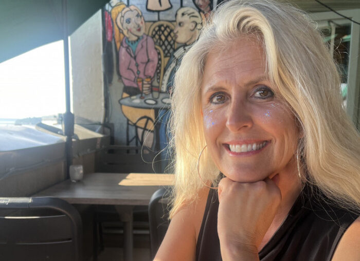 Smiling blonde woman resting chin on her hand at an outdoor cafe, with colorful mural behind her and glitter on her cheeks.