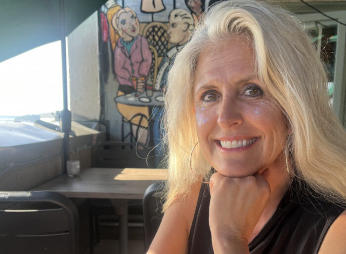 Smiling blonde woman resting chin on her hand at an outdoor cafe, with colorful mural behind her and glitter on her cheeks.