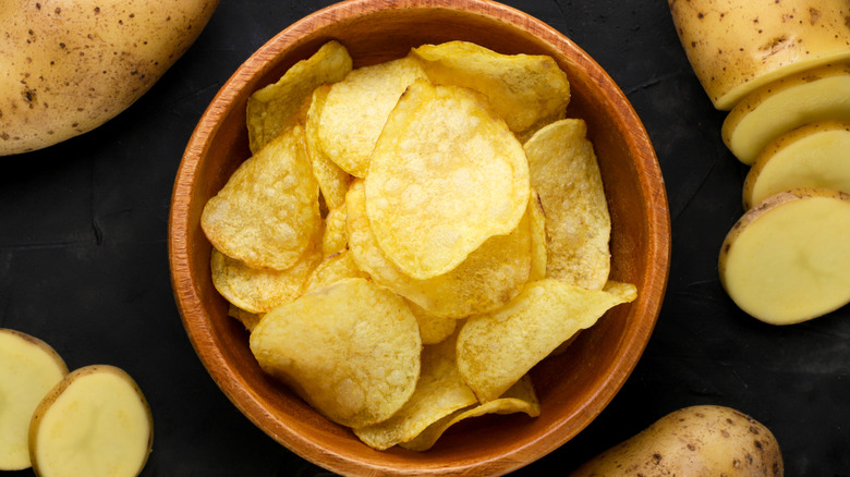 A bowl of potato chips surrounded by potatoes on a dark background