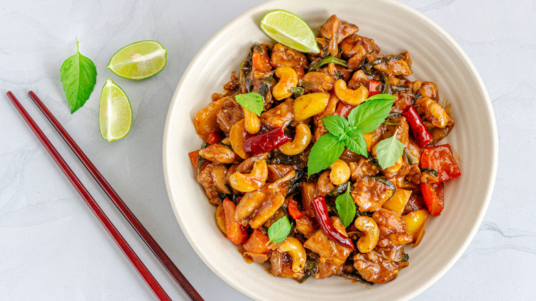 A white bowl with cashew chicken garnished with greens and a lime wedge next to chopsticks and lime